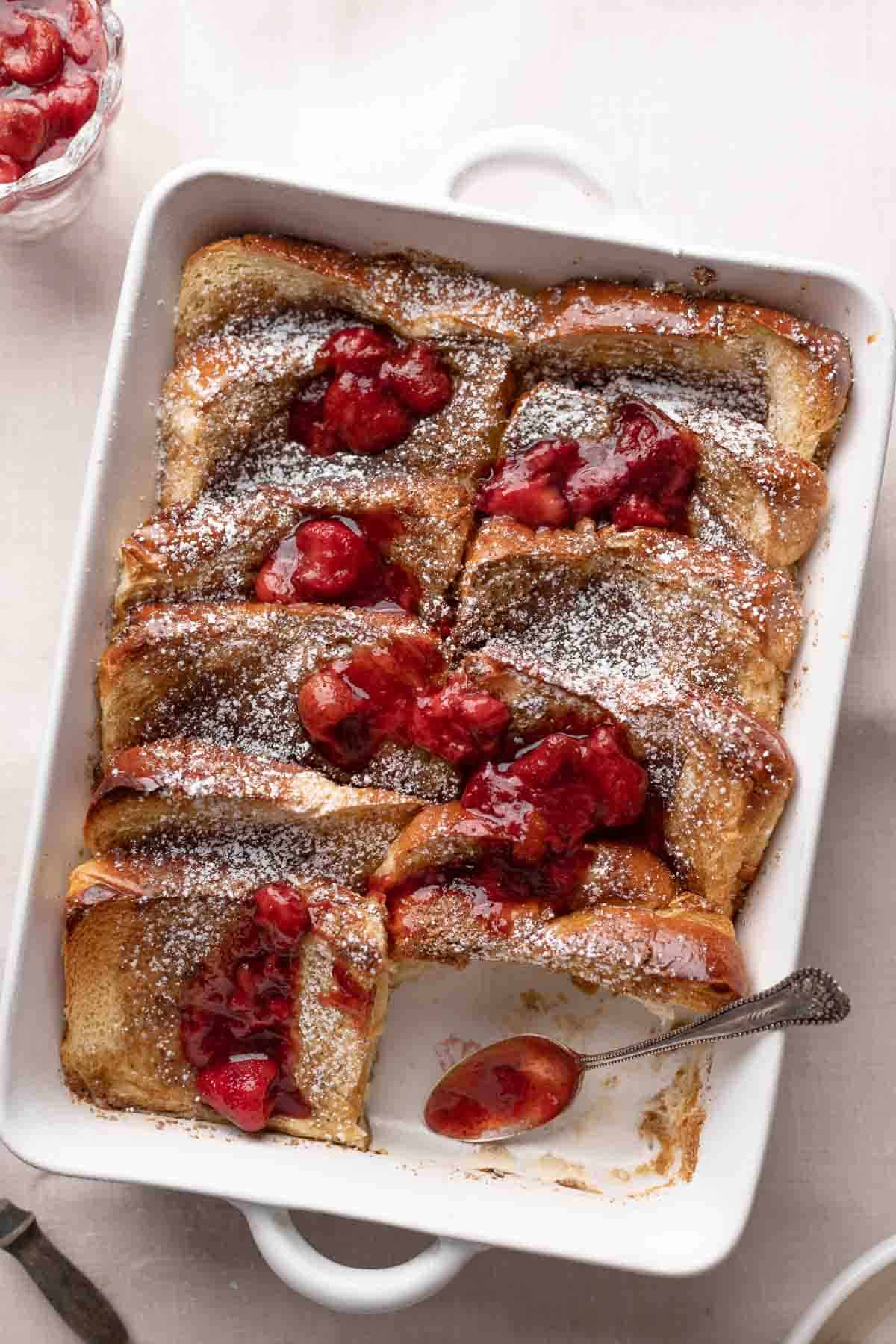 brioche french toast in a white casserole dish topped with powdered sugar and strawberries.