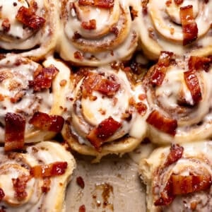 cinnamon rolls in a pan with maple frosting and candied bacon on top.