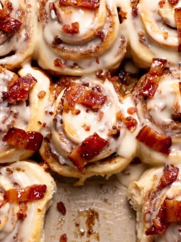 cinnamon rolls in a pan with maple frosting and candied bacon on top.