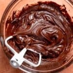 A bowl of chocolate fudge frosting with a white paddle attachment.