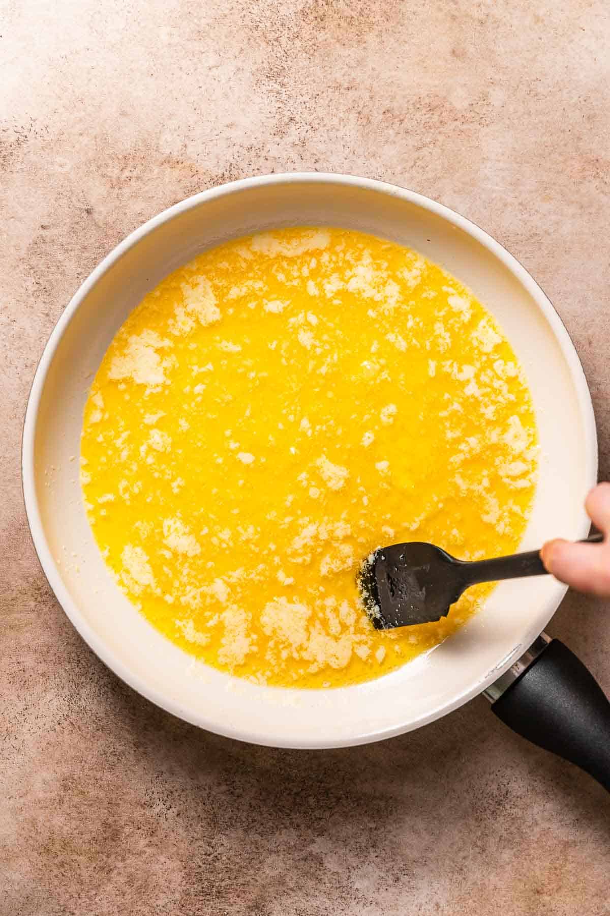 A white pan with bubbling butter being stirred with a black rubber spatula.