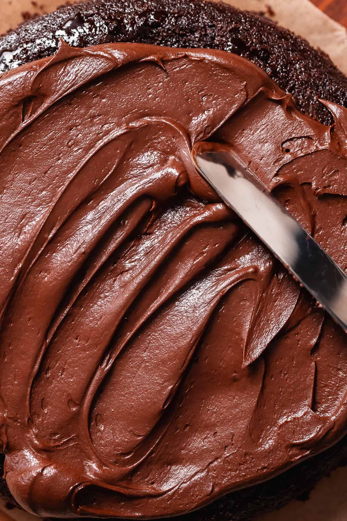 A spatula spread the chocolate buttercream frosting on a chocolate cake layer.