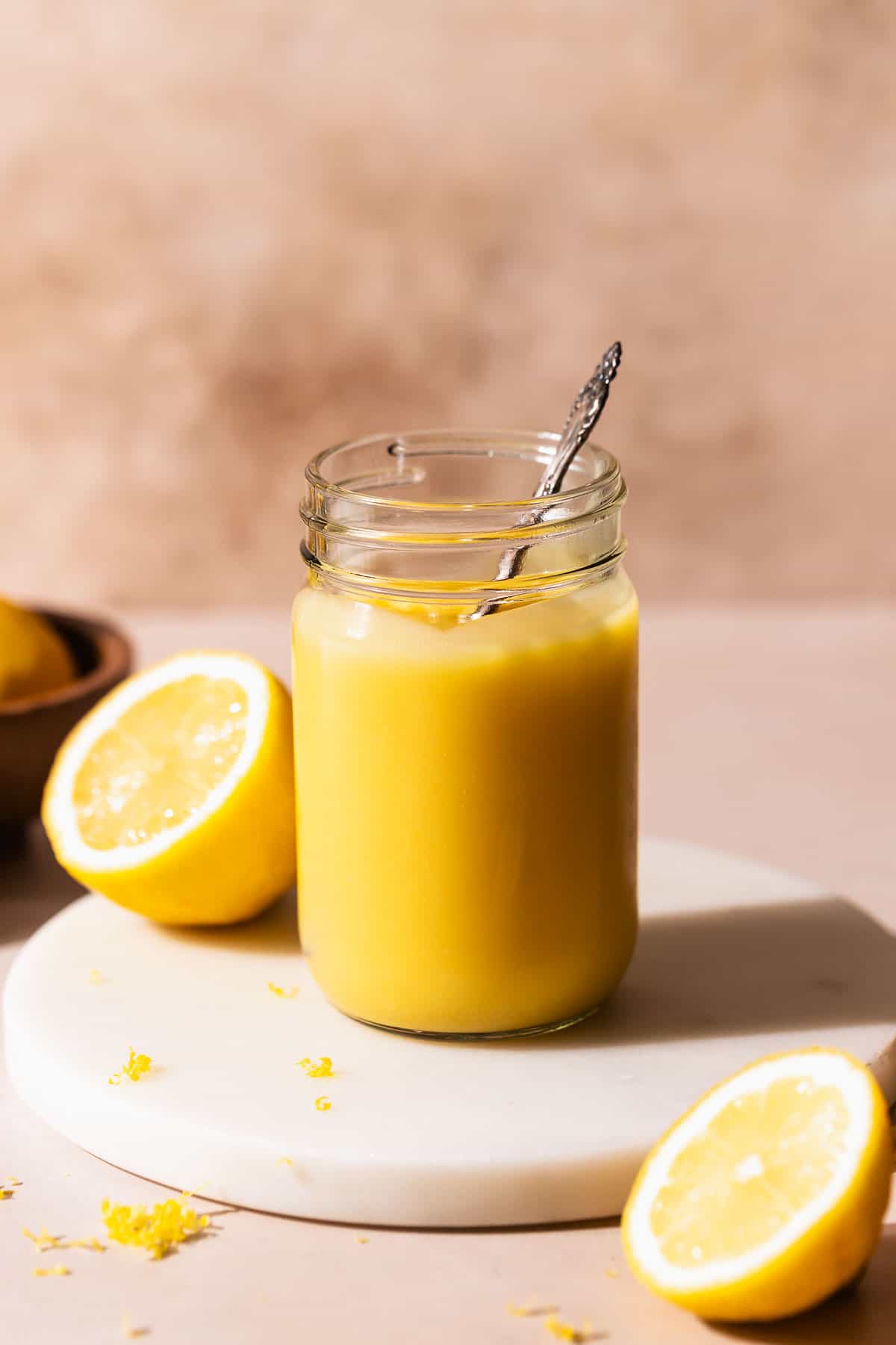 A glass mason jar of homemade lemon curd on a white marble stand with a spoon dipping into it.