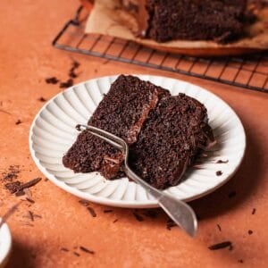A slice of moist dark chocolate cake on a white plate with a fork digging into it.