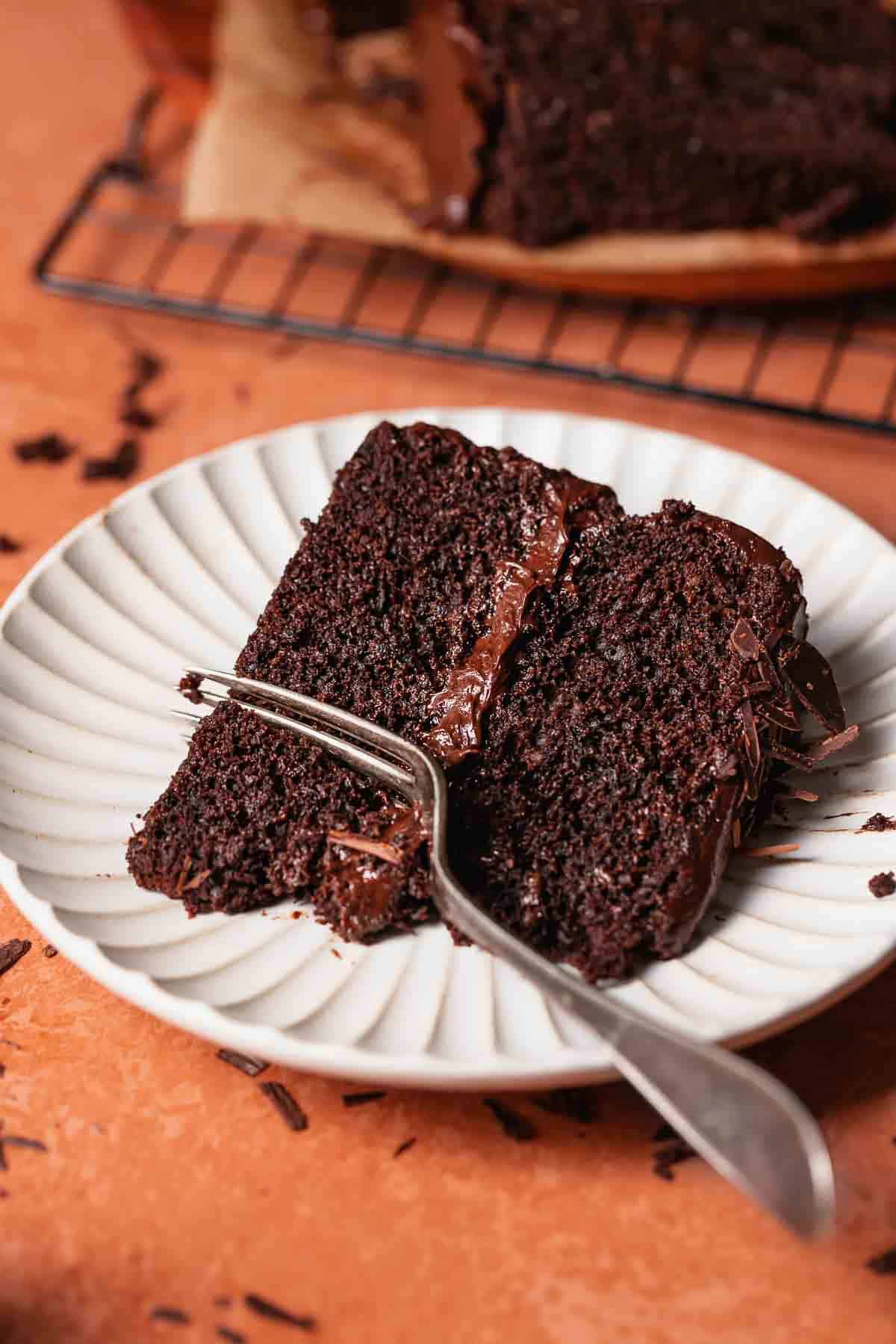 A slice of moist chocolate cake with chocolate frosting on a white plate with a fork digging into it.