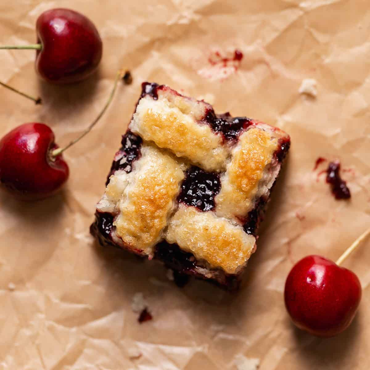 A cherry pie bar on brown parchment paper surrounded by fresh cherries.