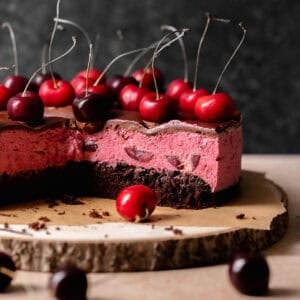 Chocolate and cherry cake on a wooden cake platter with fresh cherries on top.