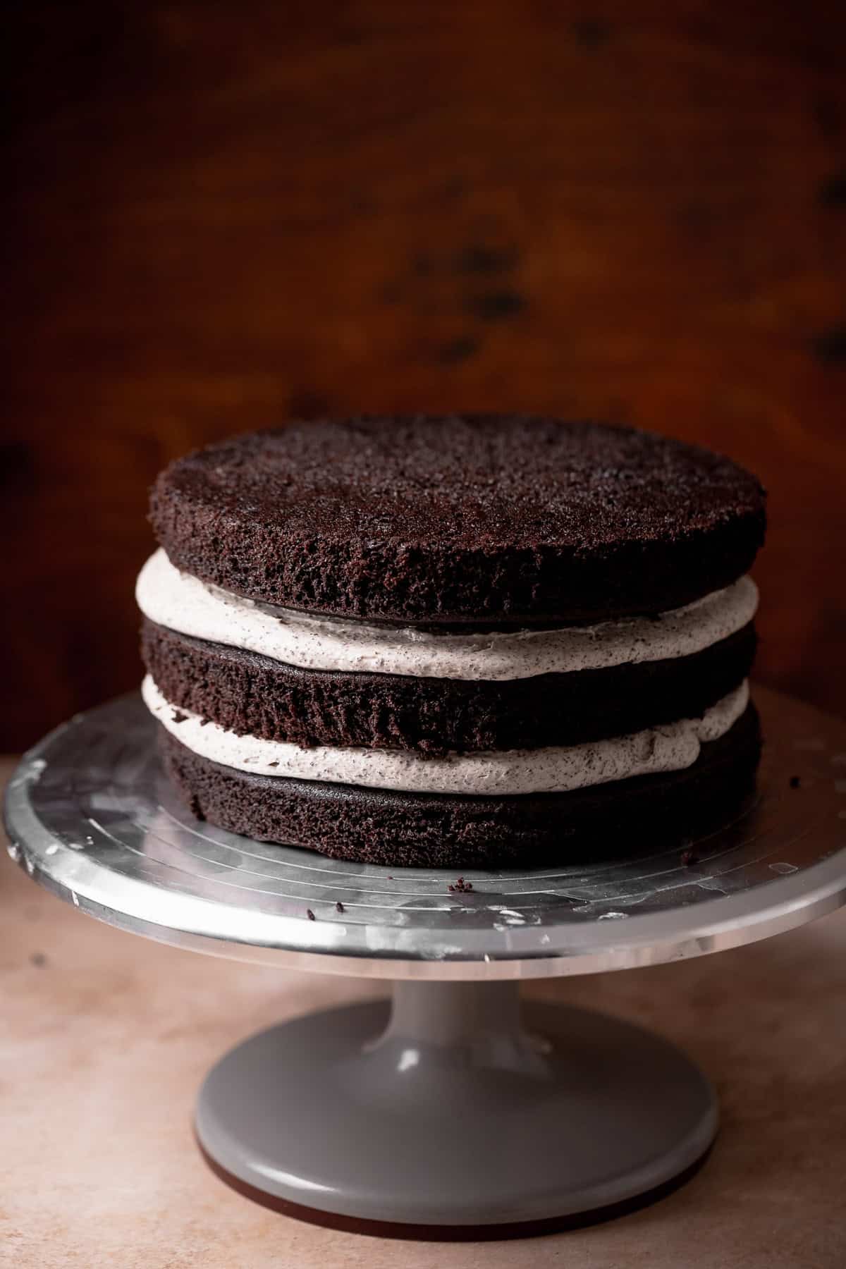 Three layers of chocolate cake with Oreo frosting in between layers on a cake stand.