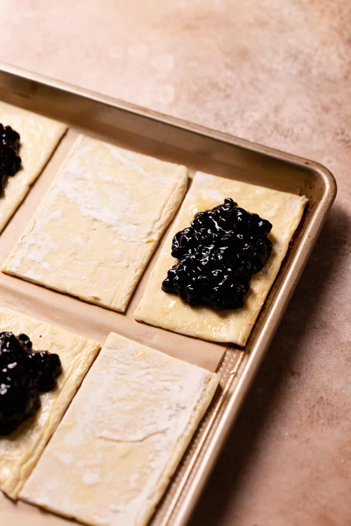 Squares of puff pastry on a baking sheet with half filled with blueberry jam.