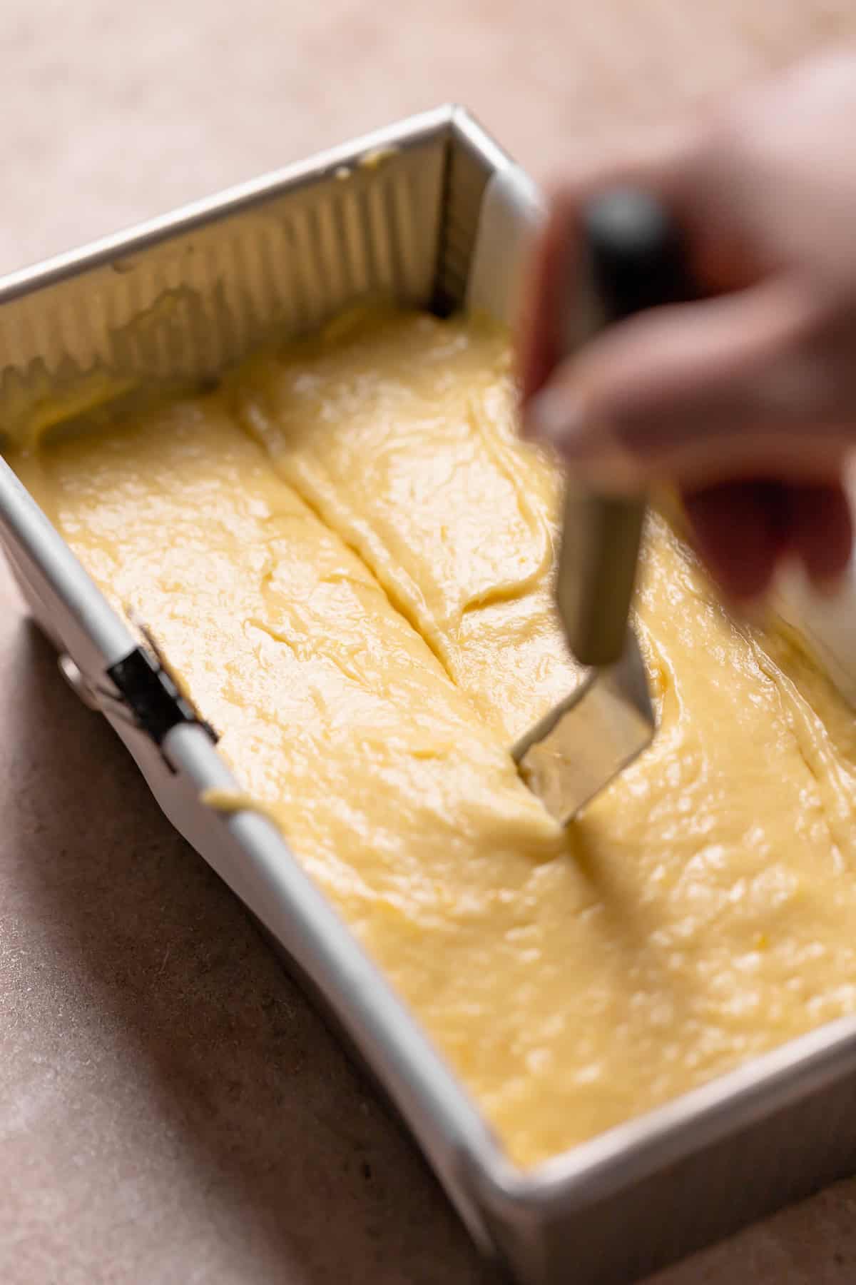 A hand dragging a knife through the center of a loaf of cake batter.