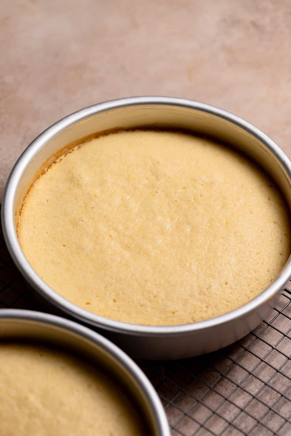 A cake pan with the baked lemon cake layer on a wire cooling rack.