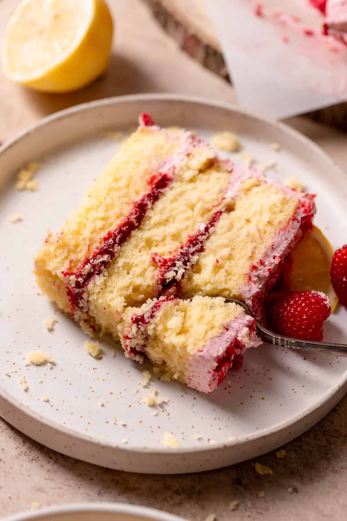 A slice of raspberry cake on a white speckled plate with a fork digging in.