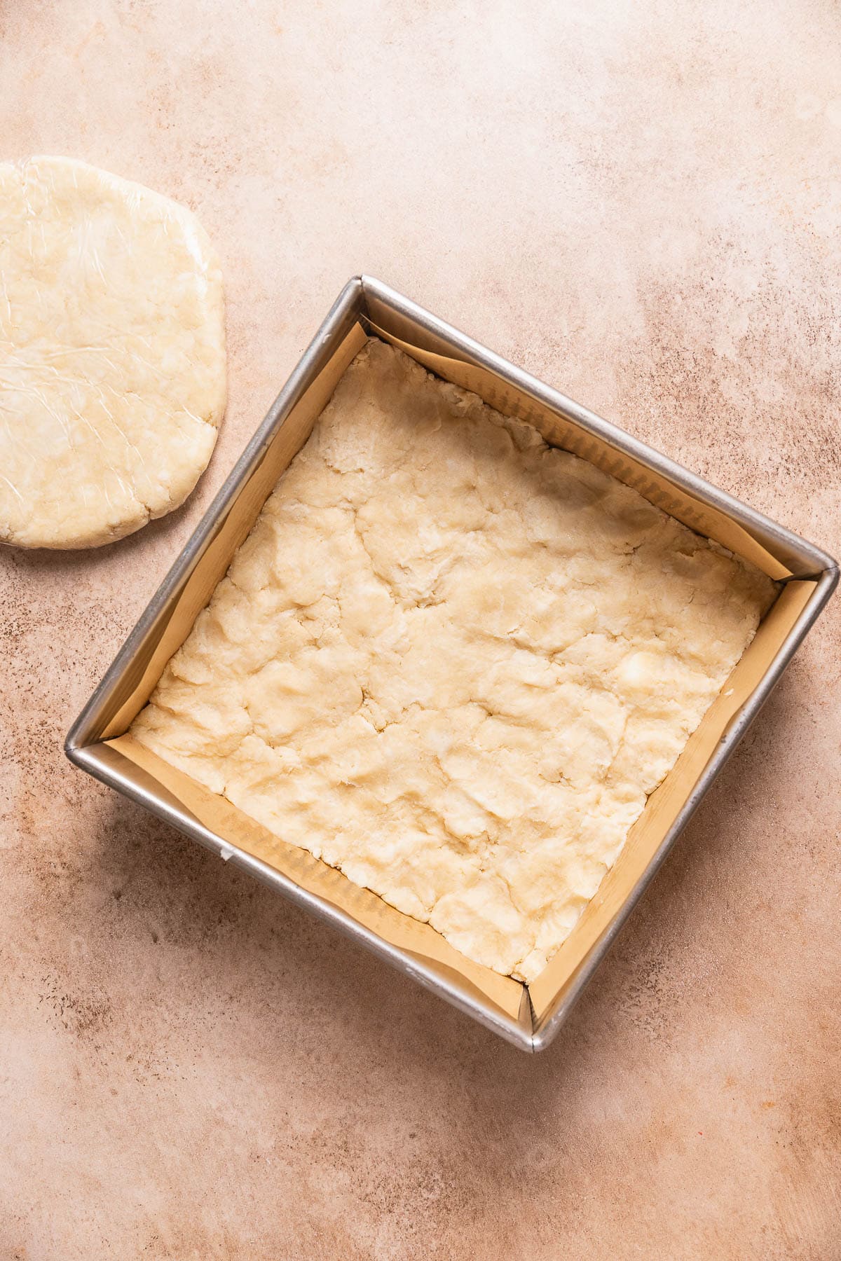 A square baking pan with pie dough pressed into the bottom and a slab of pie dough next to each other.