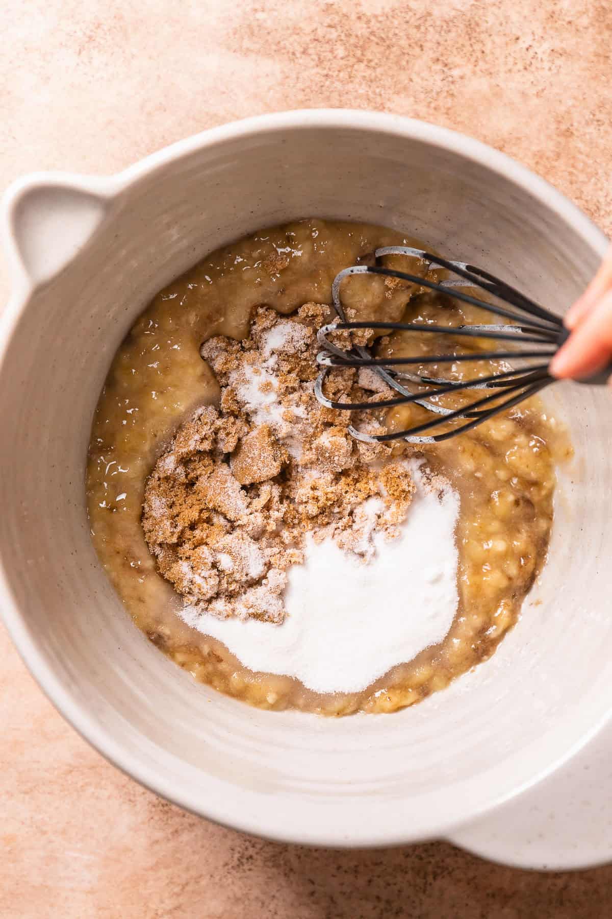 A hand whisking the sugar into the ripe mashed bananas.
