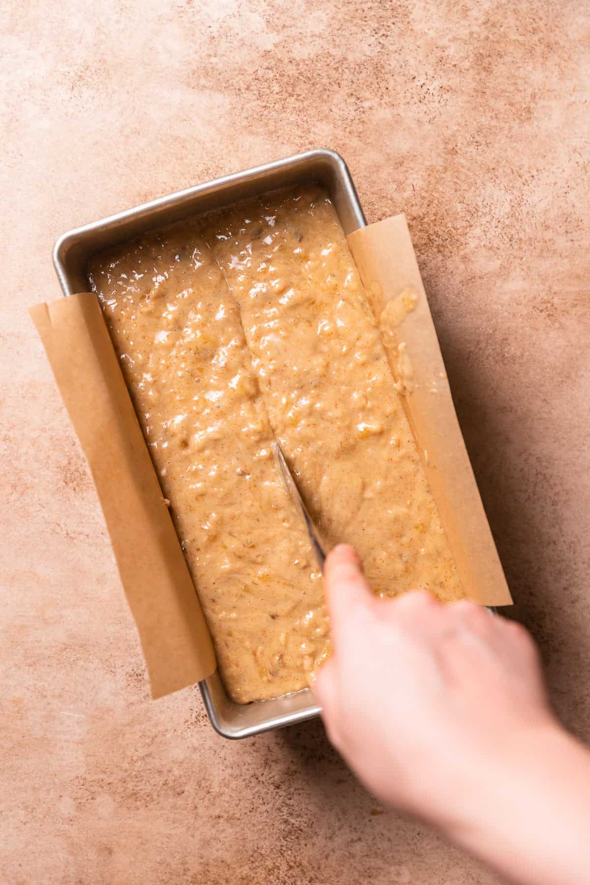 A hand dragging a butter knife through the center of the batter in a loaf pan.