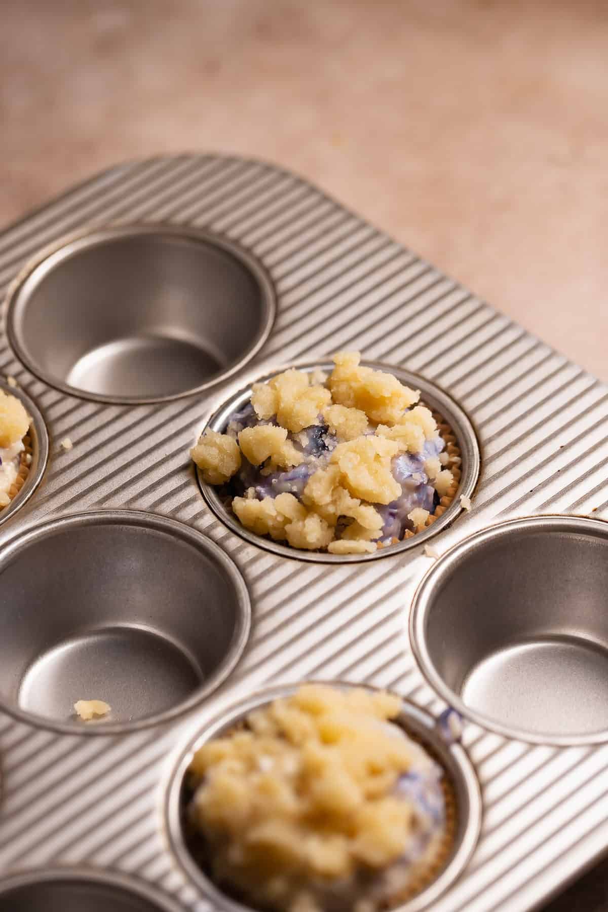 Blueberry muffin batter in a muffin pan topped with crumbs.