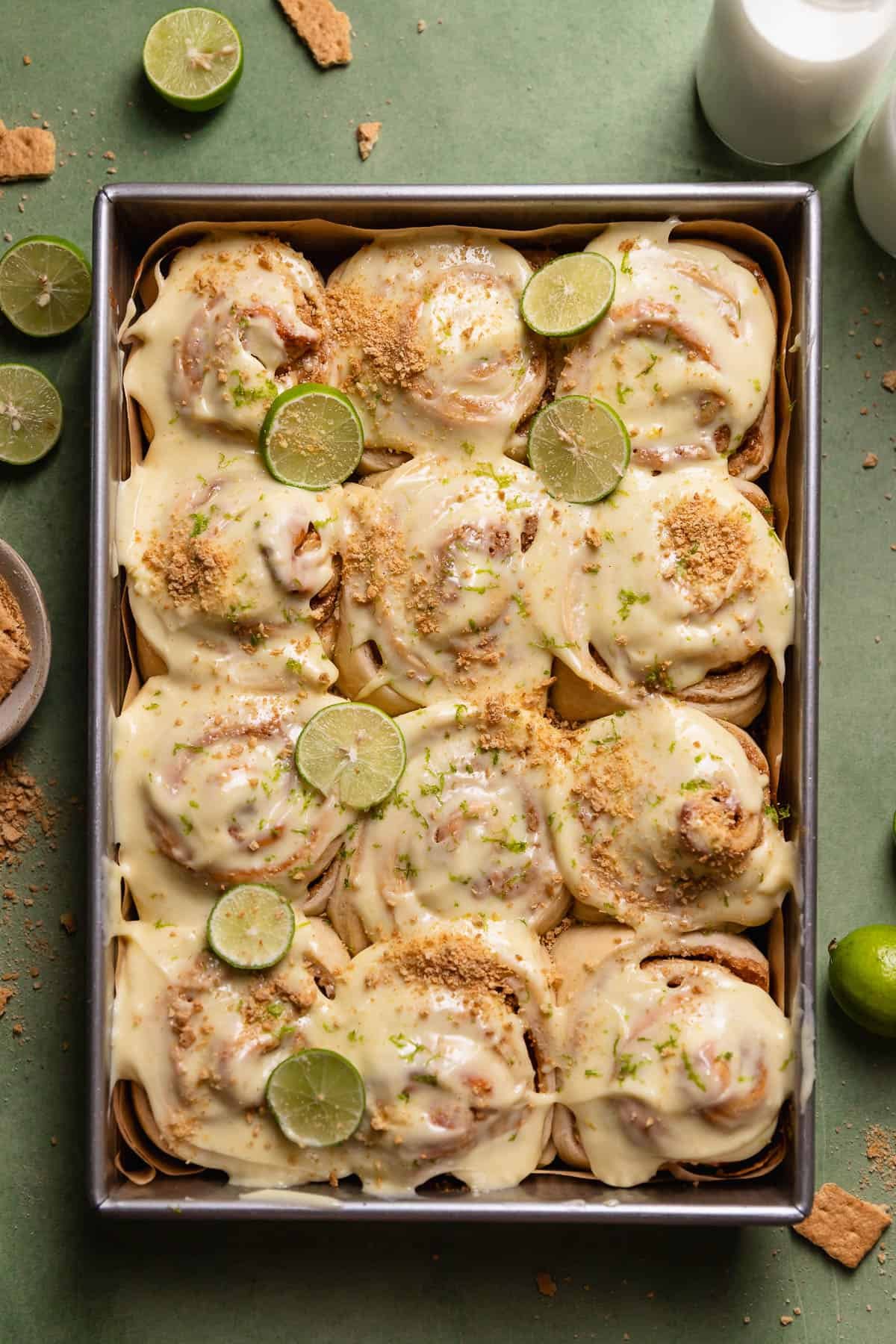 Key lime pie cinnamon rolls in a baking pan with key limes and graham cracker crumbs on top.