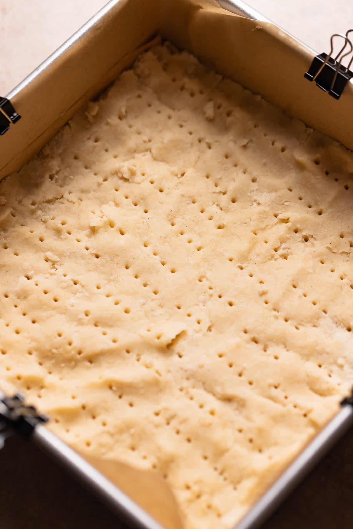 Shortbread dough pressed into a square pan pricked with a fork before baking.