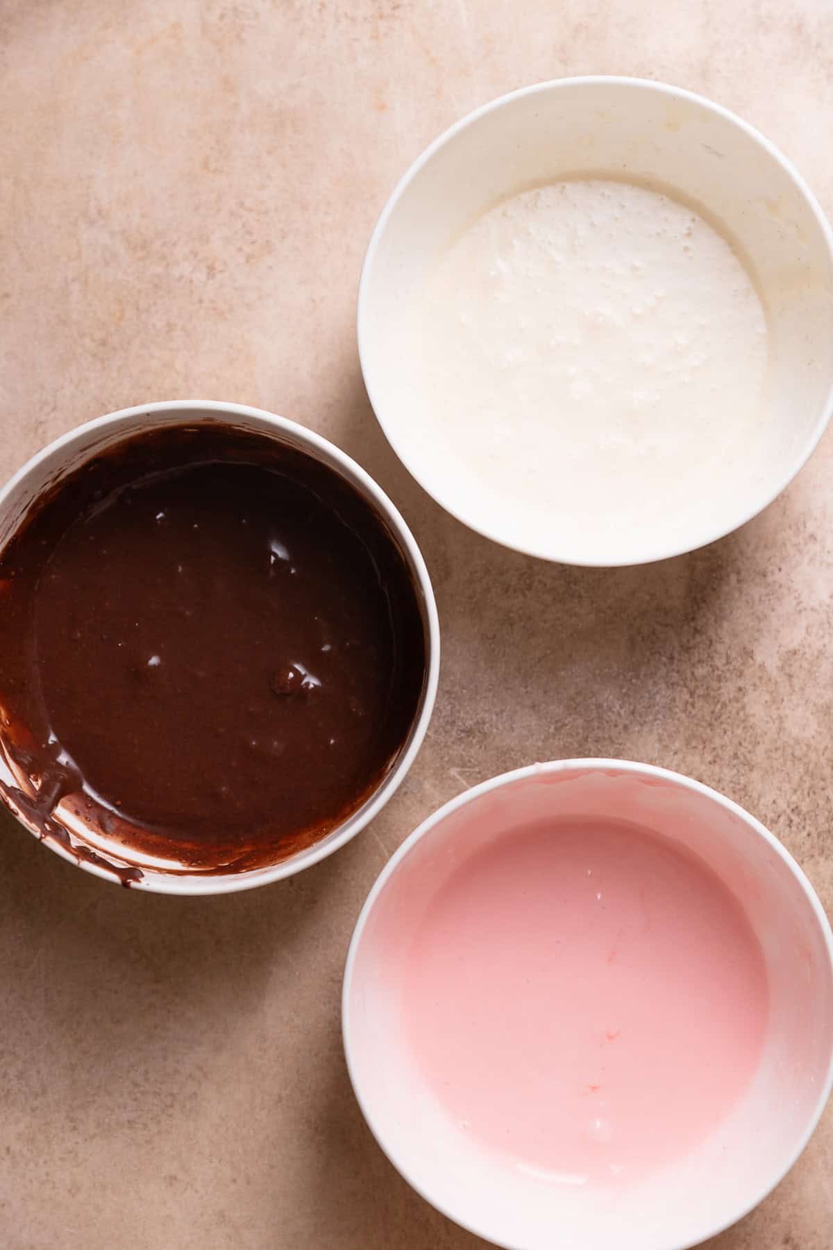 Three bowls of icing with chocolate, strawberry, and vanilla flavors.