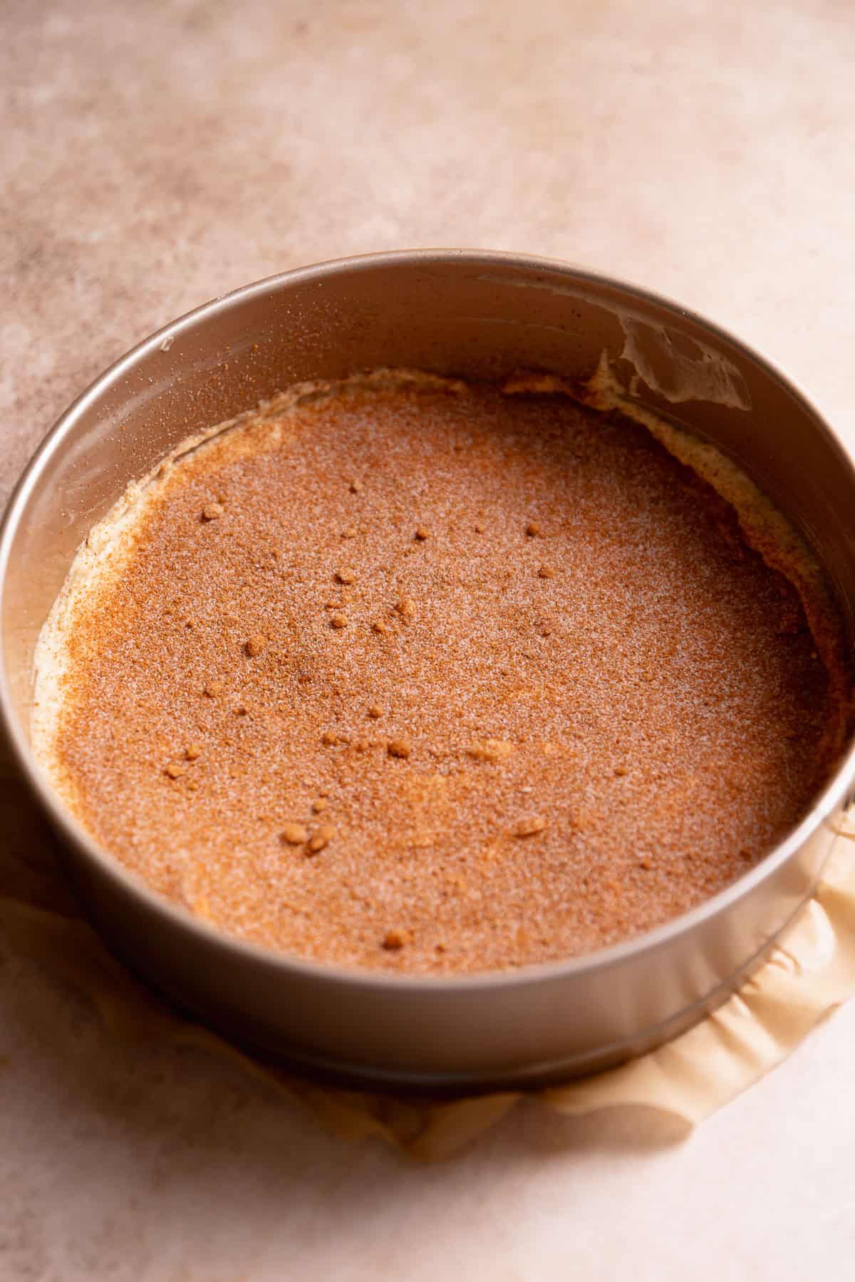 A layer of coffee cake batter in a round baking pan with cinnamon sugar sprinkled on top.