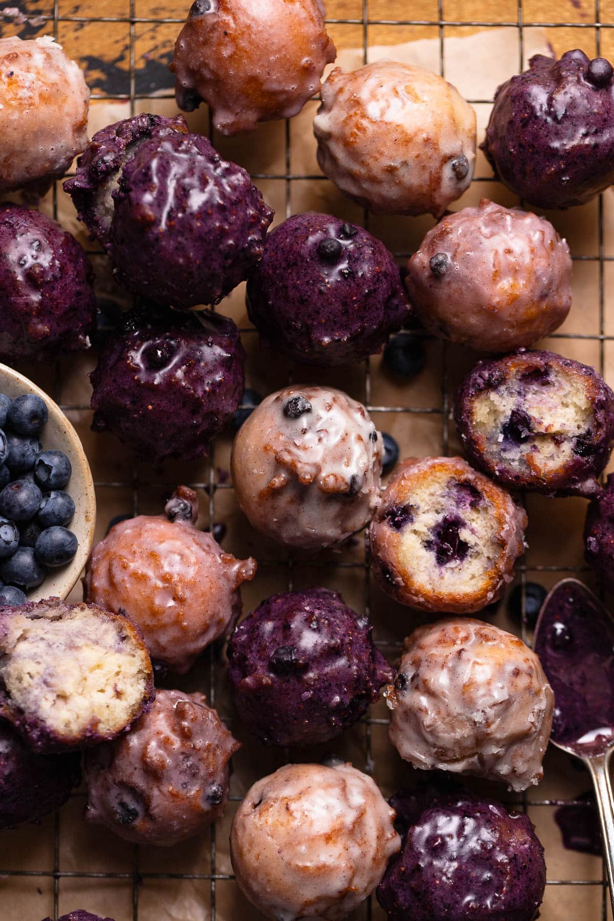 Mini blueberry cake donuts with blueberry glaze sitting in a pile on a wire rack.