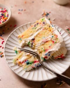A slice of confetti cake on a white plate with a fork cutting out a bite.