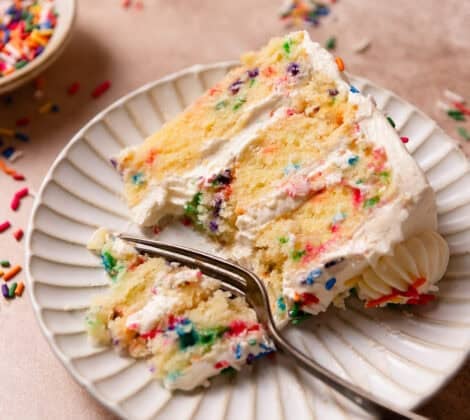 A slice of confetti cake on a white plate with a fork cutting out a bite.