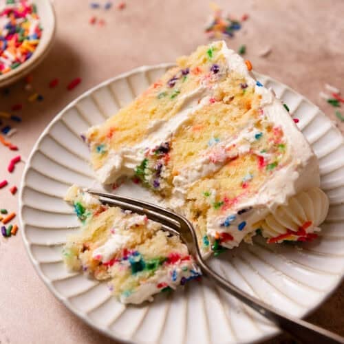 A slice of confetti cake on a white plate with a fork cutting out a bite.