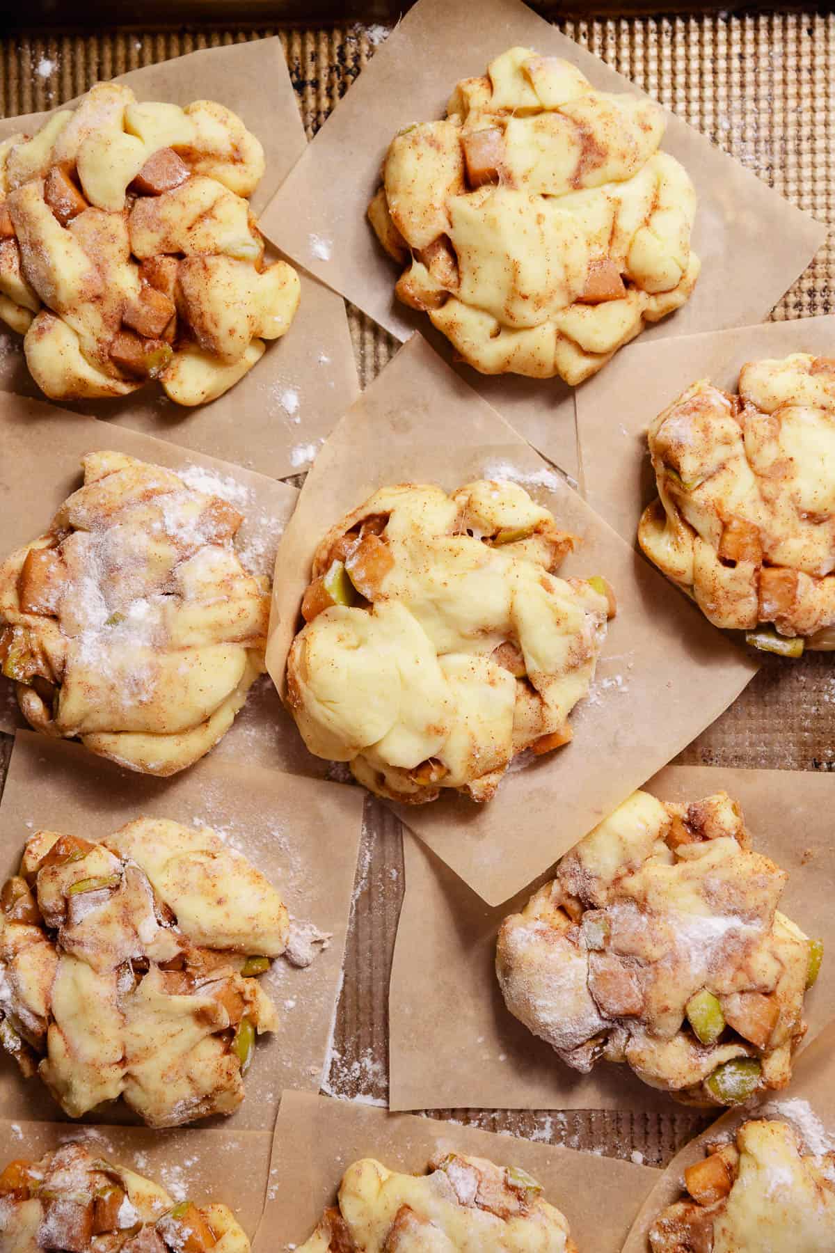 Shaped apple fritters on individual parchment squares on a baking sheet sprinkled with a bit of flour.
