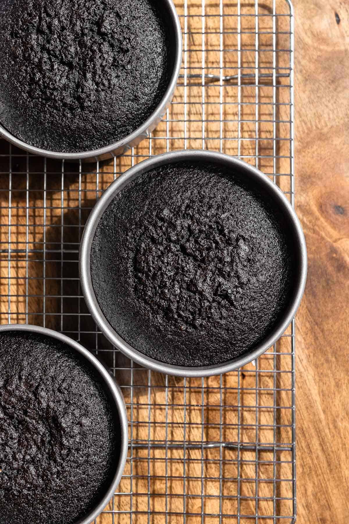 Three black chocolate cake layers in cake pans cooling on a wire rack.