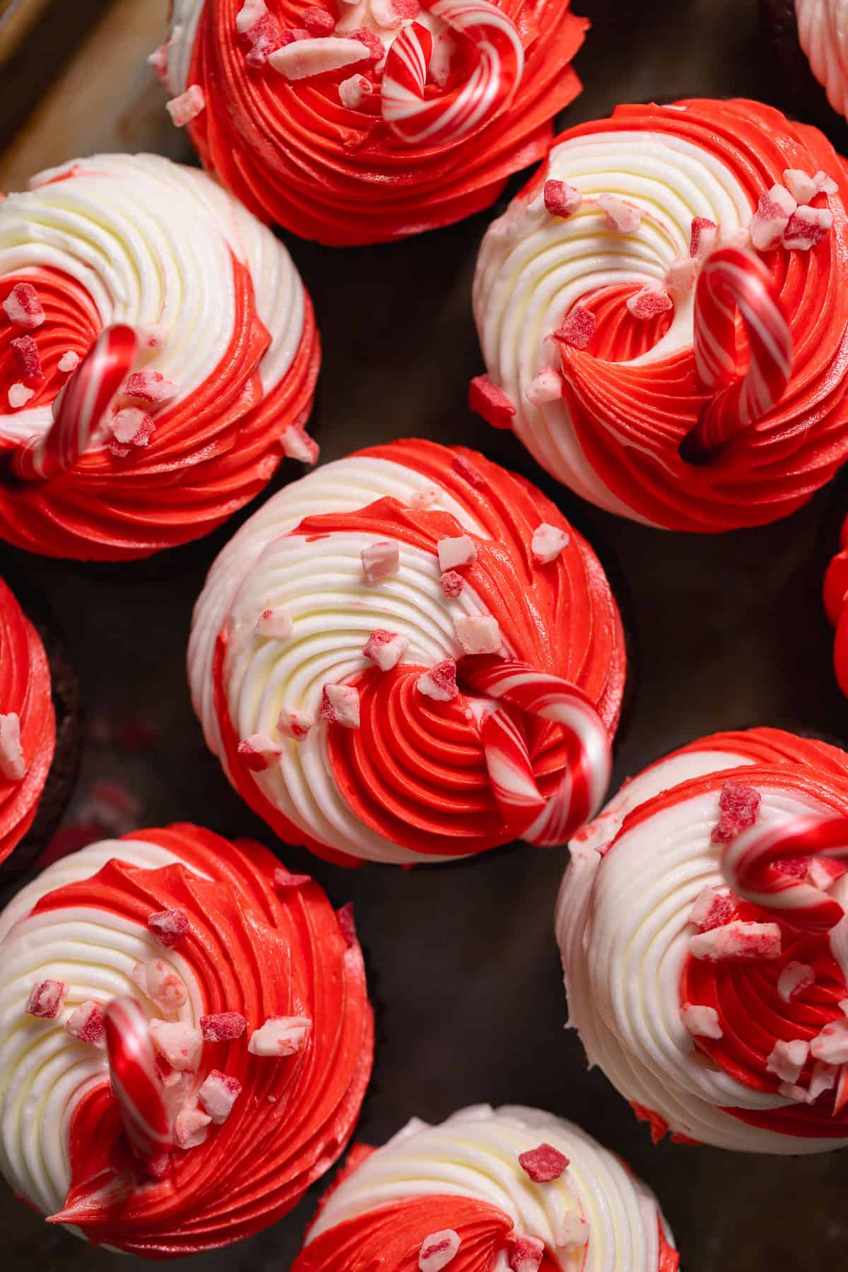 An overhead shot of red and white swirled peppermint frosting on top of chocolate cupcakes with peppermint baking chips and candy canes on top.