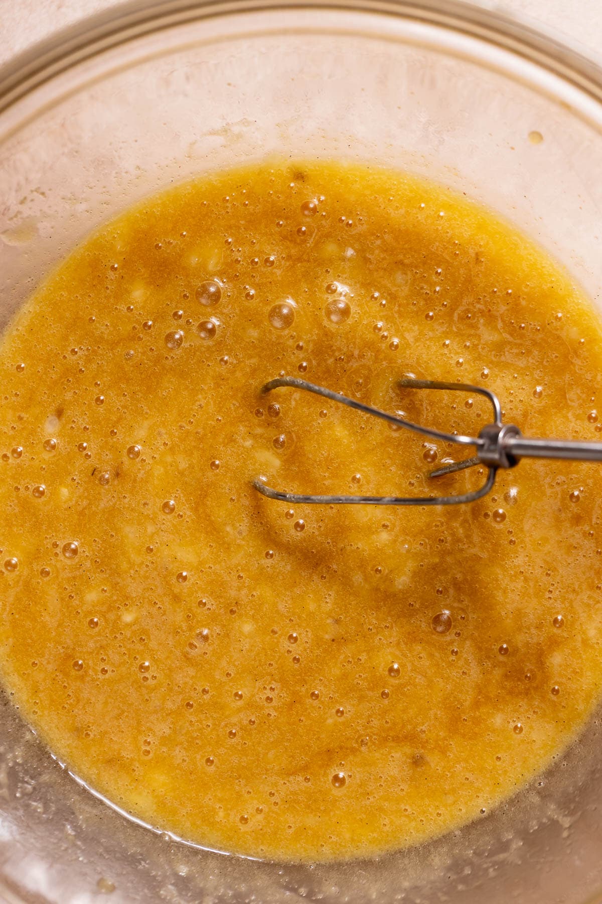 A glass bowl holds a mixture of mashed ripe bananas, melted butter, and sugar, lightly whisked together.