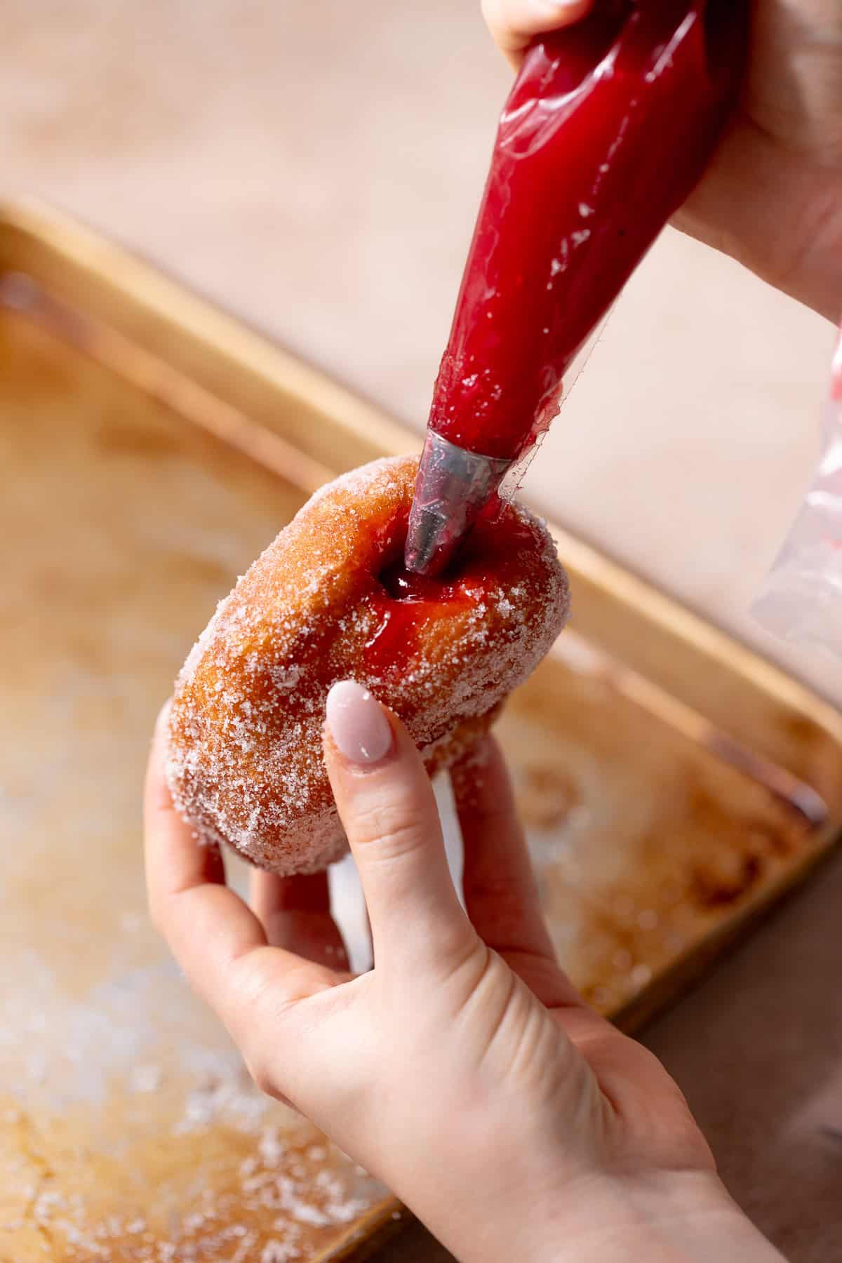 A hand with a piping bag filing a sugar-coated donut with raspberry jelly.