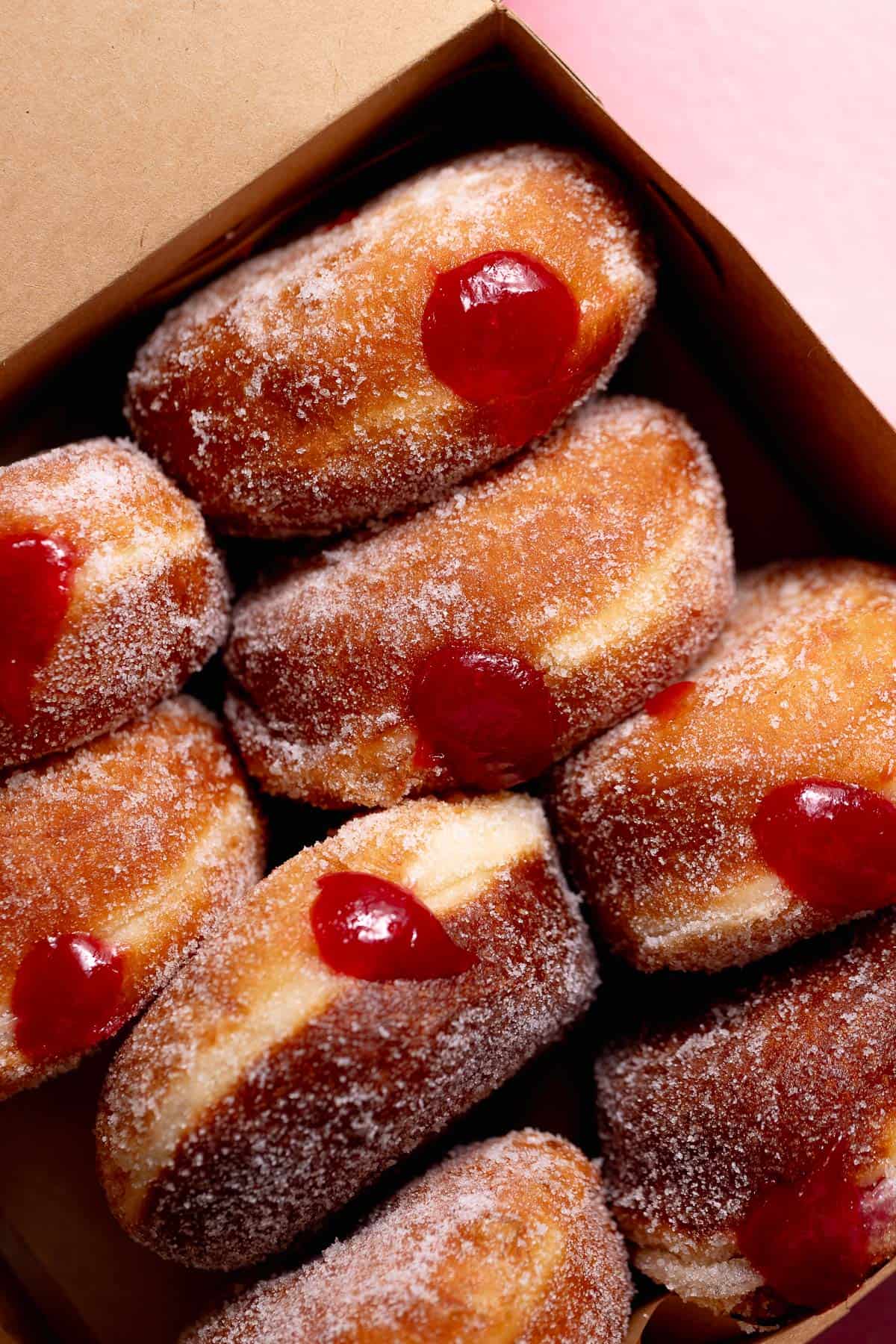 A bakery-style box filled with sugar-coated raspberry jelly donuts, each finished with a visible dollop of bright red filling.