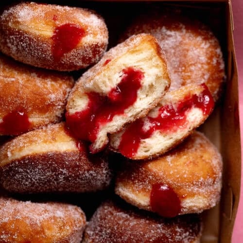A box filled with sugar-coated raspberry jelly donuts, with one donut split open to reveal a fluffy interior and generous raspberry filling.