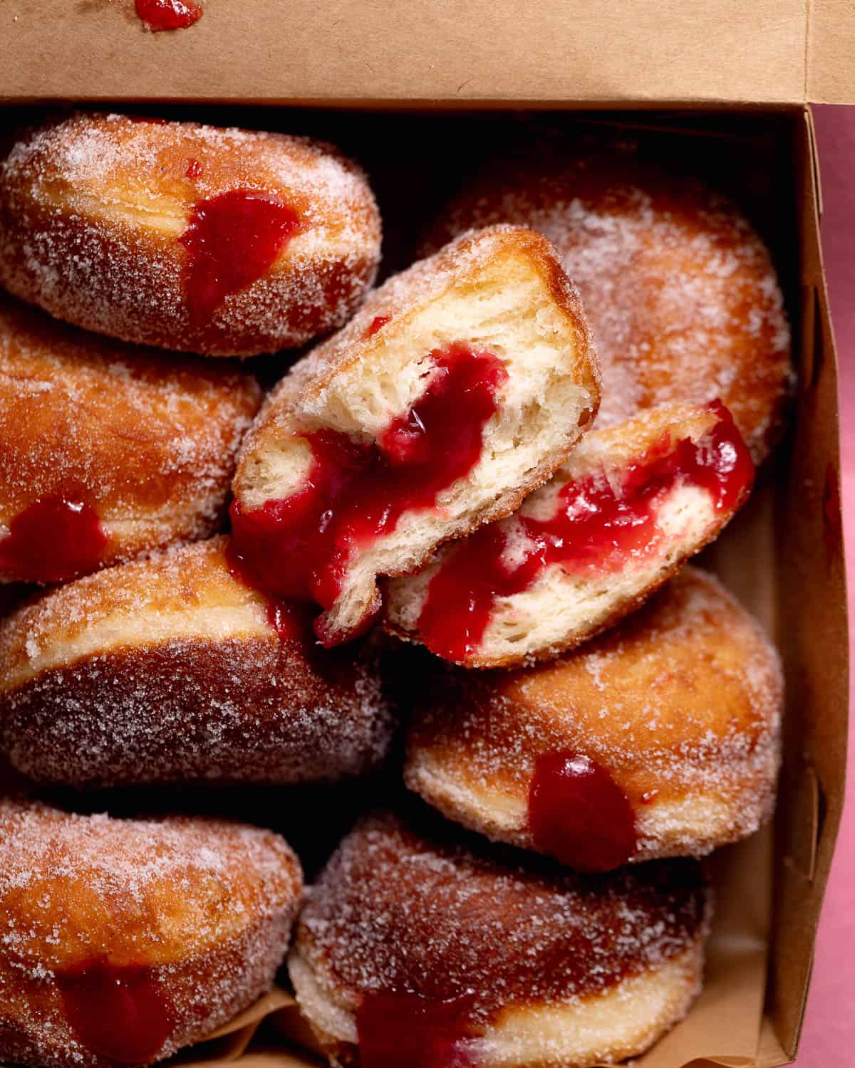 A box filled with sugar-coated raspberry jelly donuts, with one donut split open to reveal a fluffy interior and generous raspberry filling.