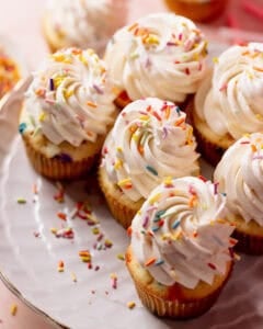 Bakery-style birthday cake cupcakes topped with tall swirls of vanilla buttercream and rainbow sprinkles on a cake stand.