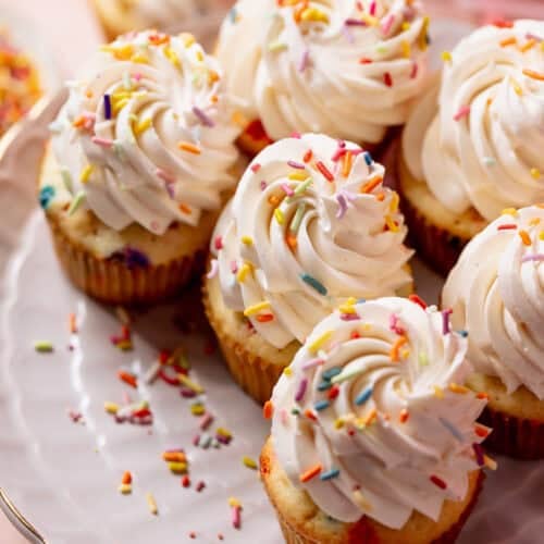 Bakery-style birthday cake cupcakes topped with tall swirls of vanilla buttercream and rainbow sprinkles on a cake stand.