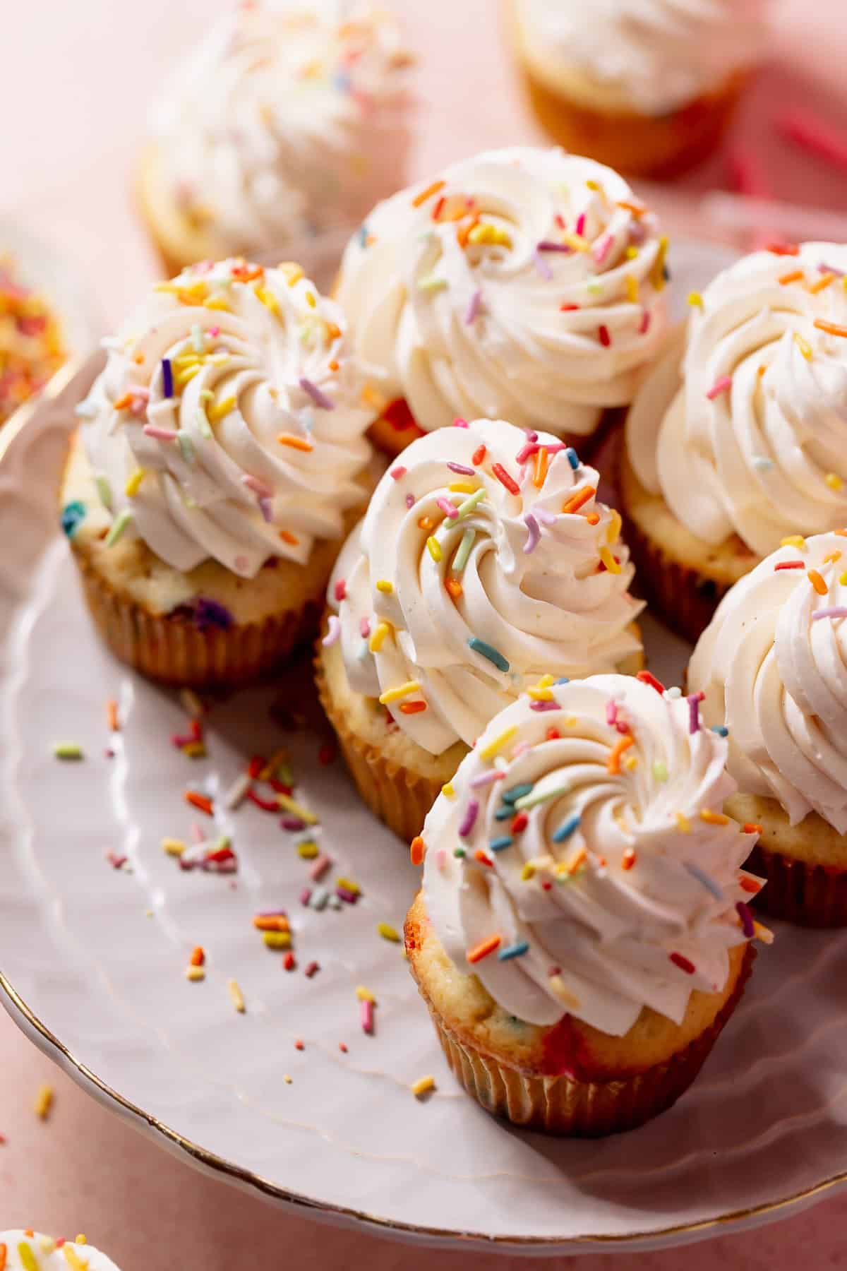 Vanilla birthday cupcakes topped with piped buttercream and rainbow sprinkles on a cake stand.