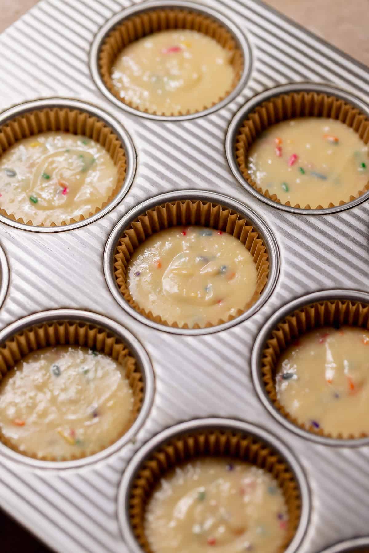 Funfetti cupcake batter spooned into lined muffin tins, speckled with rainbow sprinkles and ready to bake.