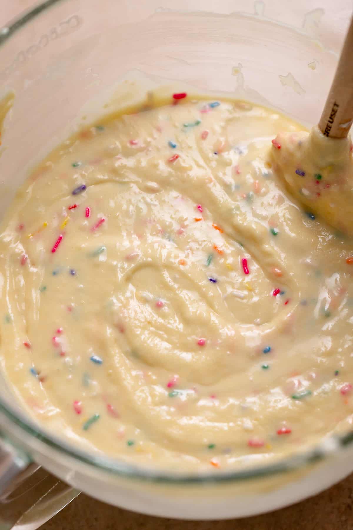 Smooth vanilla cupcake batter folded with rainbow sprinkles in a glass bowl, showing a thick, glossy texture before baking.