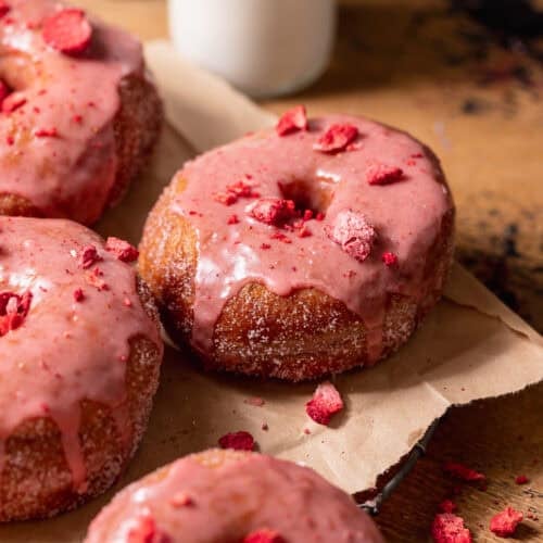 Strawberry-glazed donuts topped with freeze-dried strawberries on a cooling rack with milk in the background.