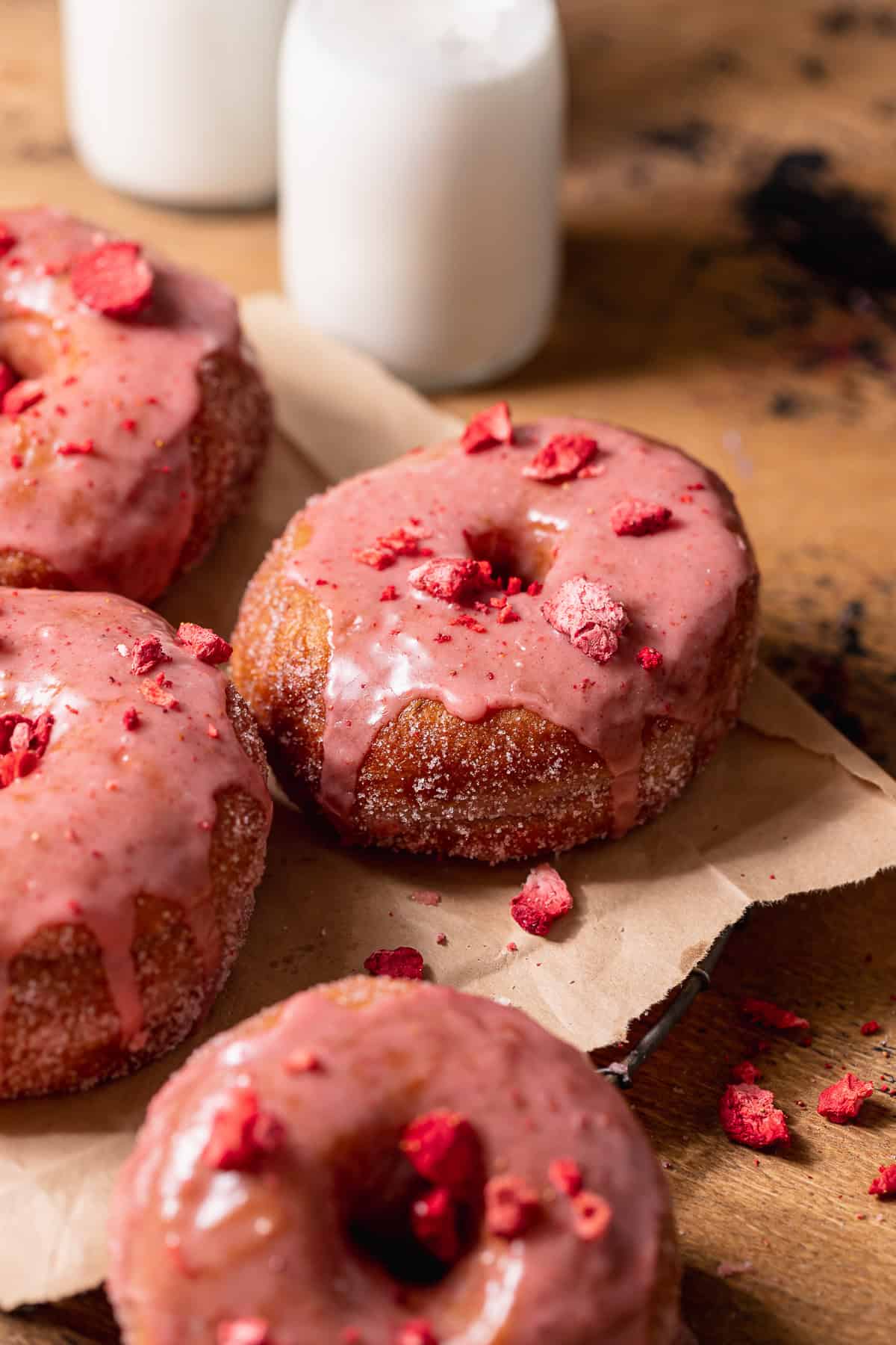Freshly fried donuts coated in pink strawberry glaze and topped with crushed freeze-dried strawberries on parchment paper.