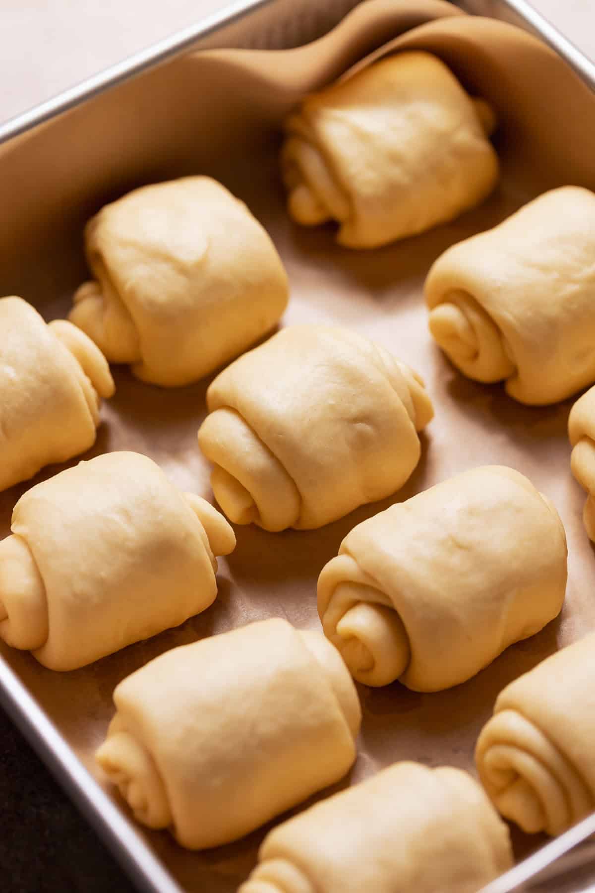 Shaped milk bread rolls spaced in a metal pan before proofing, showing folded layers.
