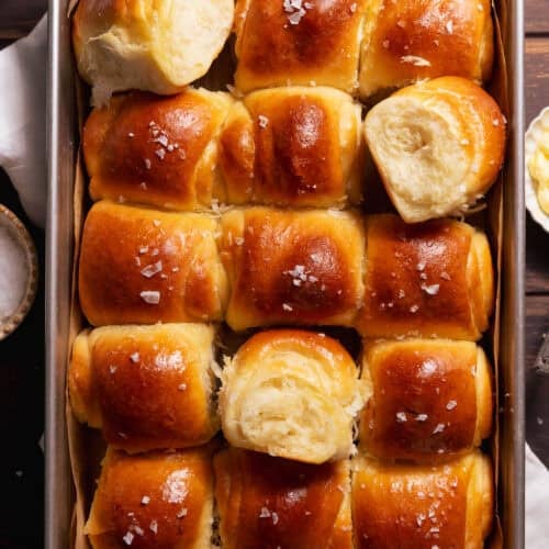Golden brown milk bread dinner rolls baked in a square pan with flaky sea salt on top.