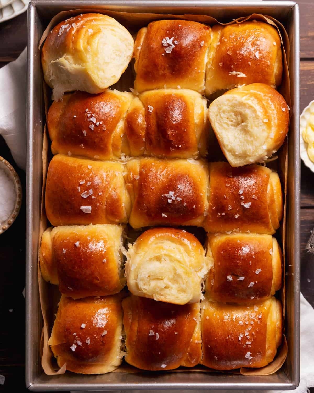 Golden brown milk bread dinner rolls baked in a square pan with flaky sea salt on top.