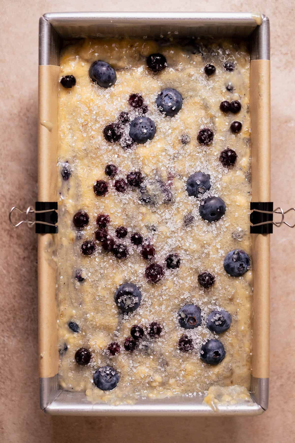 Unbaked blueberry banana bread batter in a lined loaf pan topped with sugar and berries.
