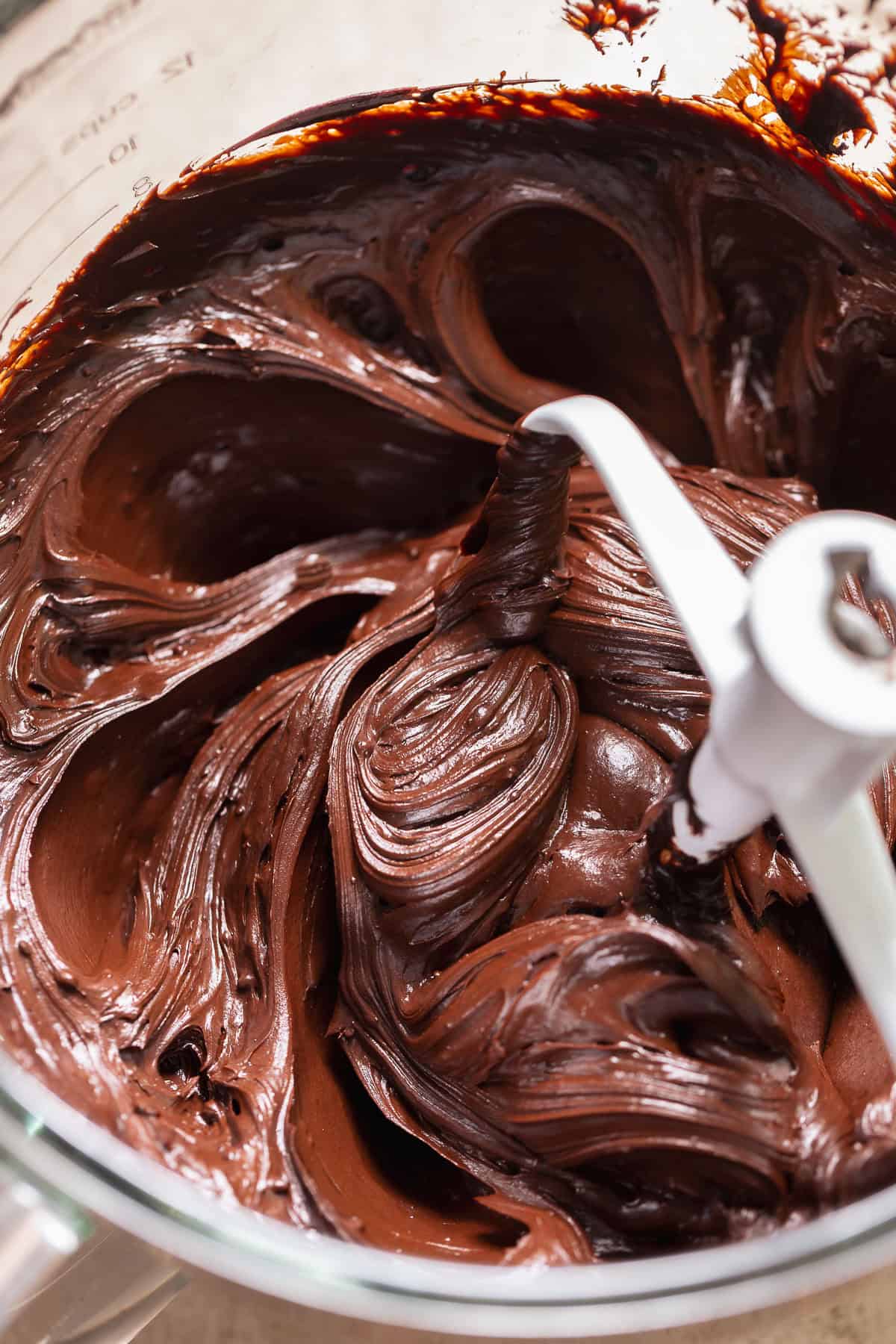 Thick chocolate fudge frosting being mixed in a bowl with a paddle attachment.