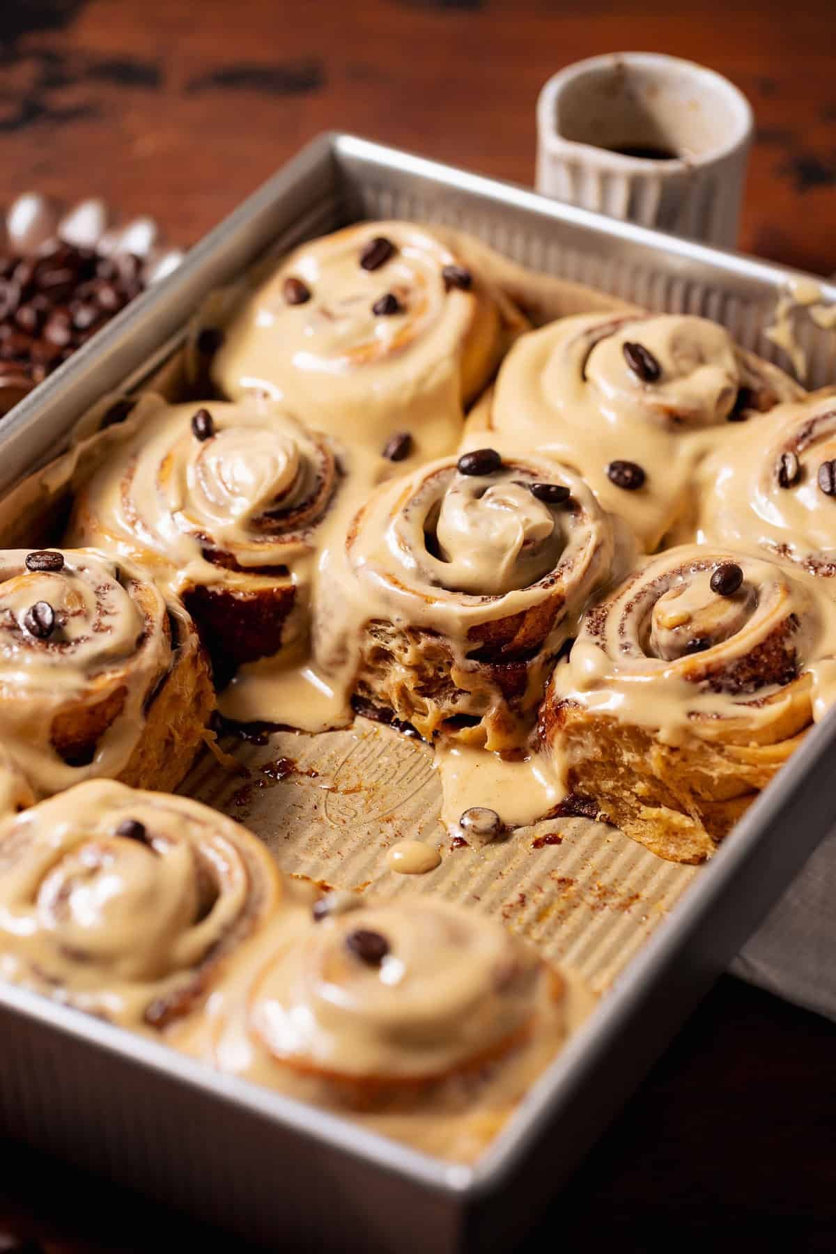 A pan of soft coffee cinnamon rolls with espresso cream cheese frosting and coffee beans on top, with a few rolls pulled out showing the gooey center.