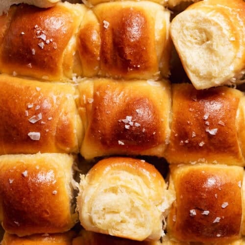 Golden brown milk bread dinner rolls baked in a square pan with flaky sea salt on top.